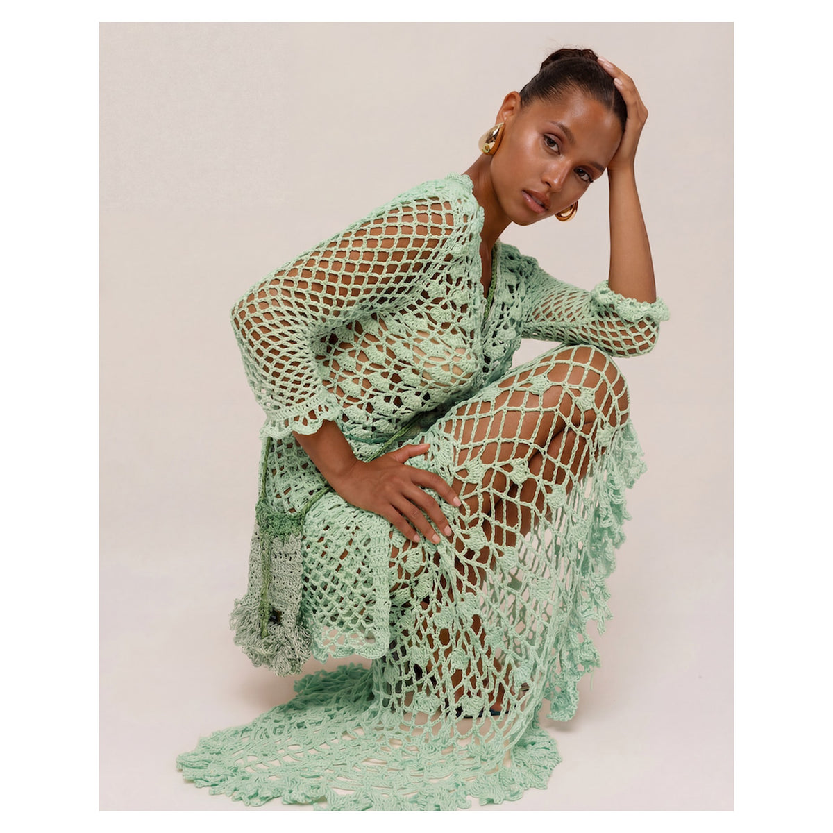 A woman kneels on one knee, wearing the ANDREEVA California Mint Handmade Knit Midi Skirt with large open-weave patterns. She pairs it with gold hoop earrings, has her hair pulled back, and poses against a plain background.