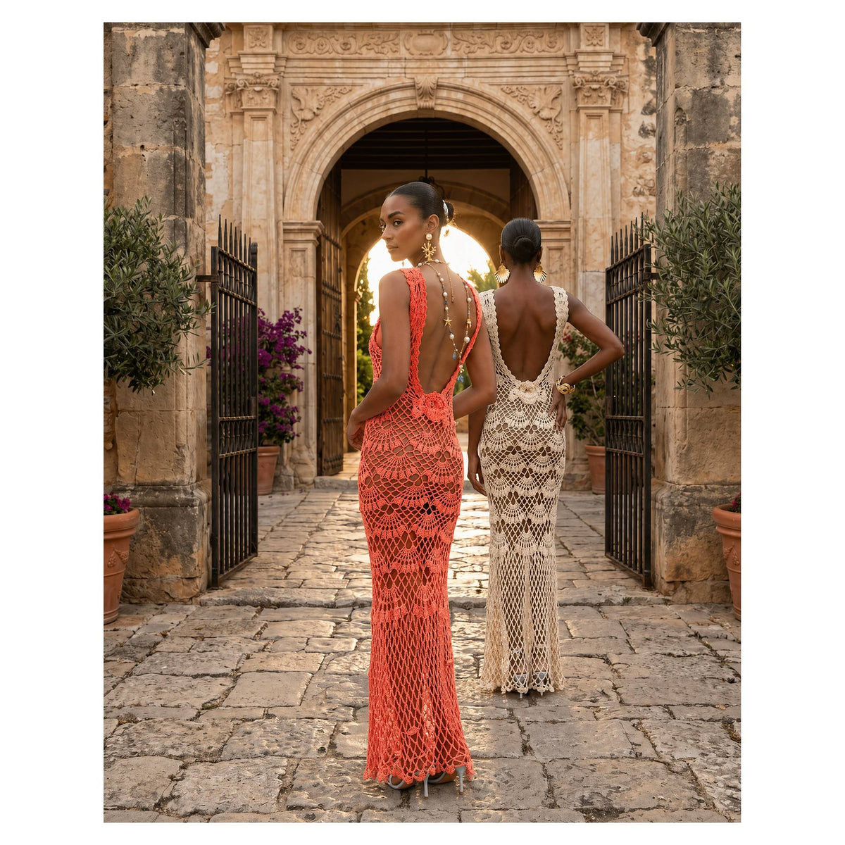 Two women in ANDREEVA handmade crochet dresses—one in the Coral Shell Maxi with pearls, the other in cream—stand on a stone path before a grand archway, radiating coastal elegance among lush plants and bright flowers.