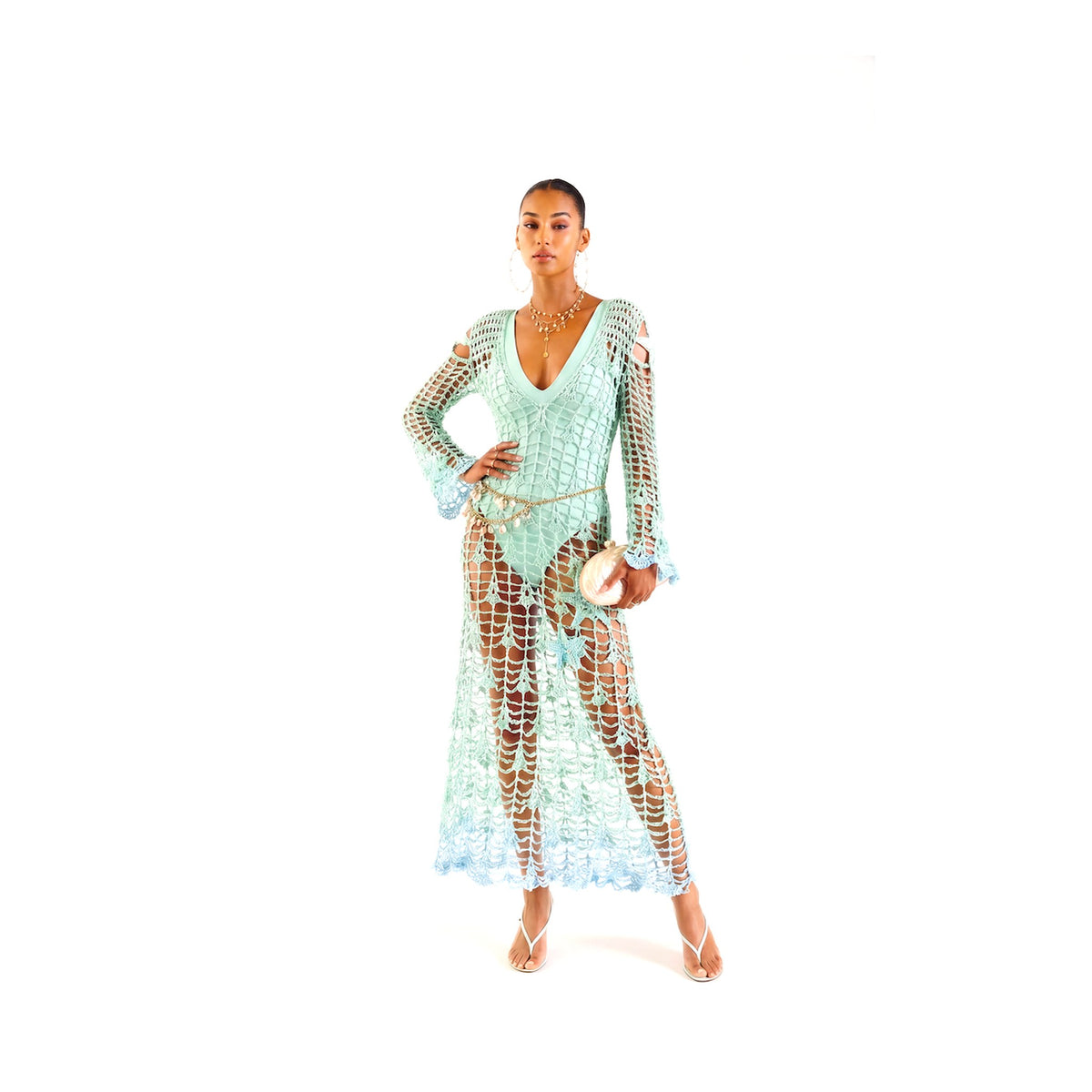A woman poses confidently in the ANDREEVA Aqua Marine Handmade Maxi Cover-Up Dress, layered over a matching bikini. She completes the look with a clutch, sandals, jewelry, and hoop earrings against a white background.
