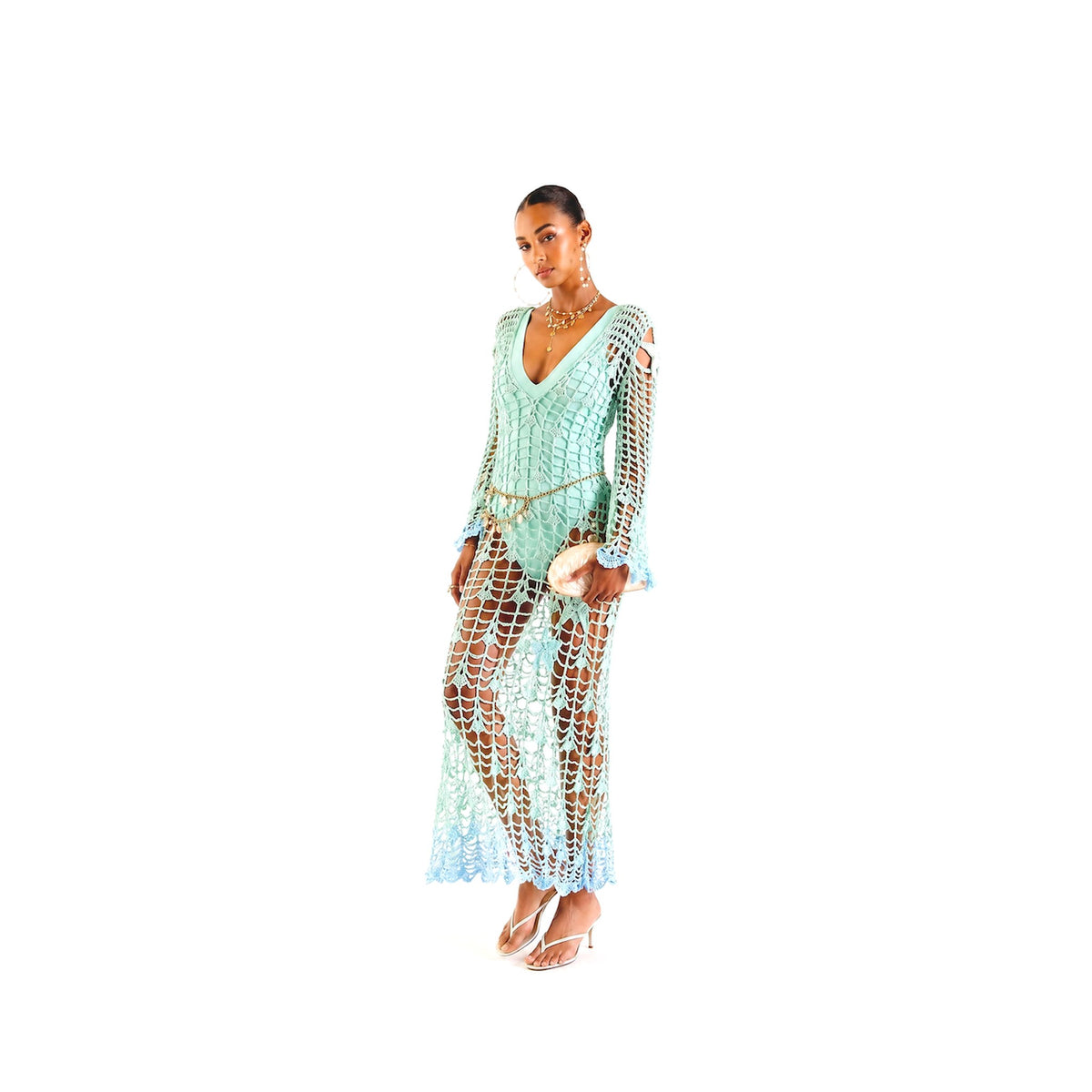 A woman poses in the ANDREEVA Aqua Marine Handmade Maxi Cover-Up Dress over a matching swimsuit, accessorized with a small clutch, strappy sandals, and gold jewelry, against a white background.