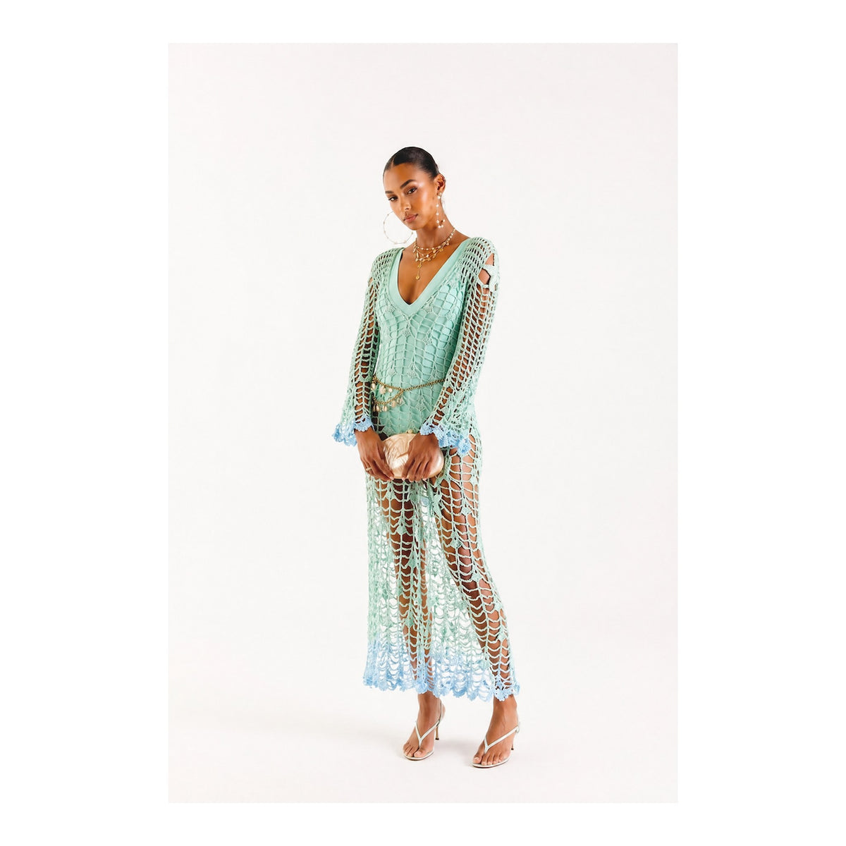 A woman models the ANDREEVA Aqua Marine Handmade Maxi Cover-Up Dress in mint green Brazilian cotton, featuring a sheer crochet design, V-neck, and long sleeves. She pairs it with a tan clutch, strappy sandals, and layered necklaces.