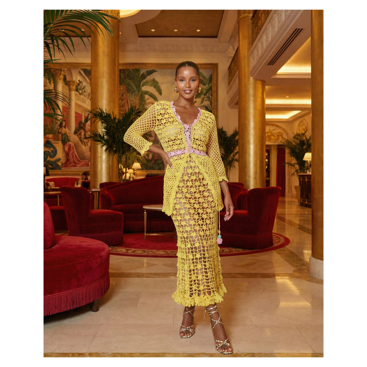 A woman stands in an elegant lobby, wearing the ANDREEVA California Yellow Handmade Crochet Shirt over a lavender undergarment with strappy heels. The setting includes golden columns, red velvet furniture, and lush plants.