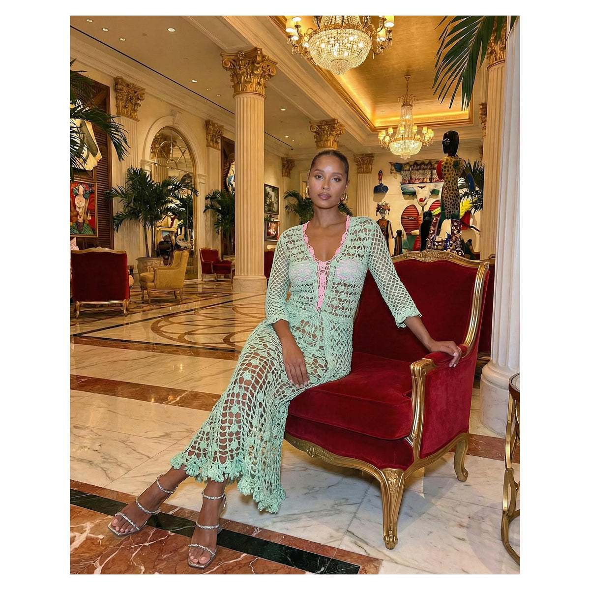 A woman in the ANDREEVA California Mint Handmade Crochet Shirt sits on a red and gold armchair in a marble-floored room with columns, chandeliers, statues, and lush greenery, embodying effortless boho charm.