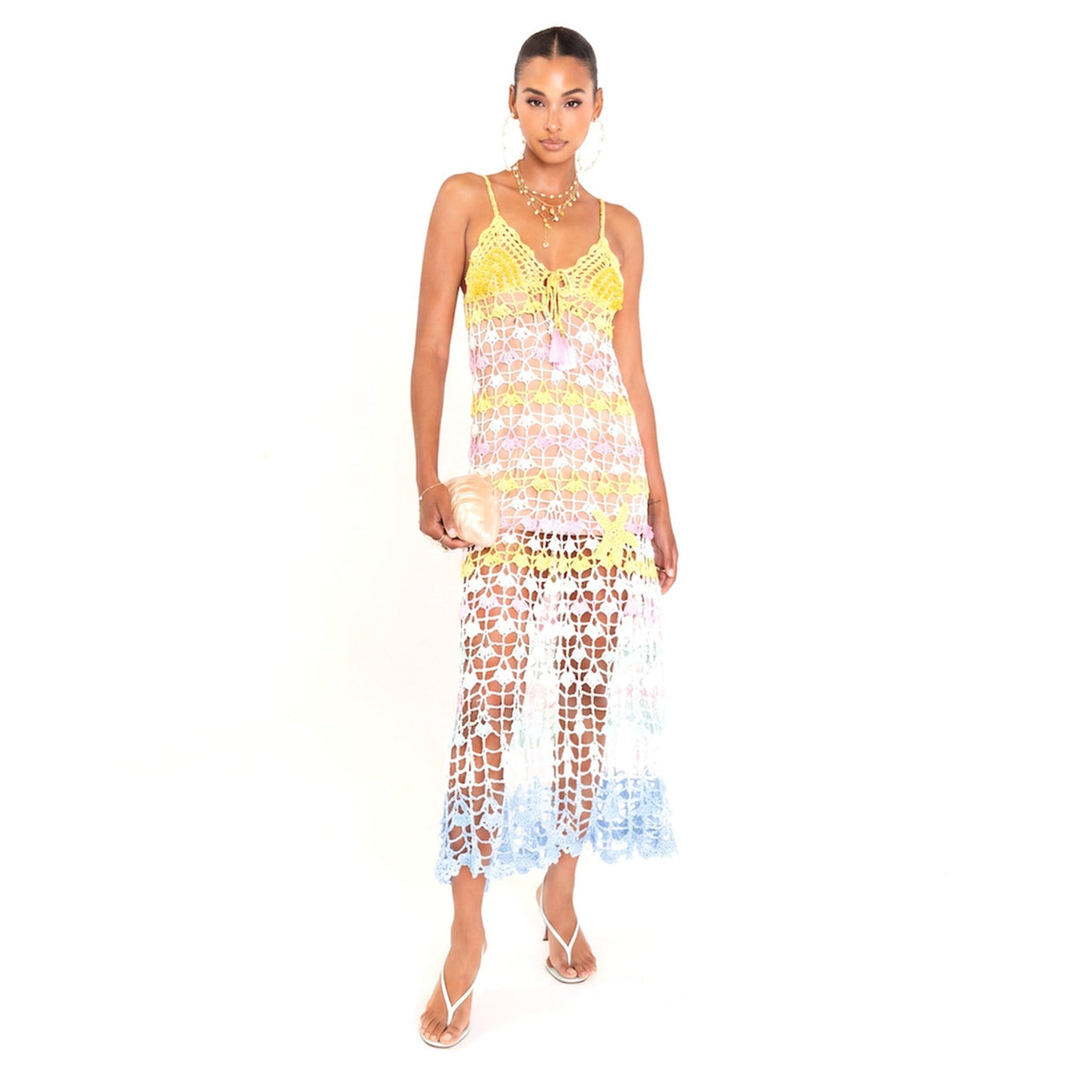 A woman models the ANDREEVA Flower Marine Handmade Maxi Crochet Dress, crafted from mercerized Brazilian cotton with yellow, pink, and blue floral crochet. She accessorizes with a shiny clutch, strappy heels, layered necklaces, and earrings against a white backdrop.