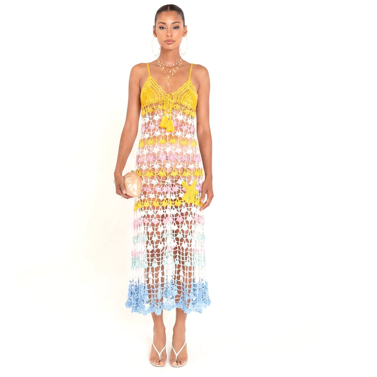 A woman models the ANDREEVA Flower Marine Handmade Maxi Crochet Dress, featuring yellow, pink, white, and blue floral crochet patterns. She holds a gold clutch, wears strappy heels, large hoop earrings, and has her hair tied back.