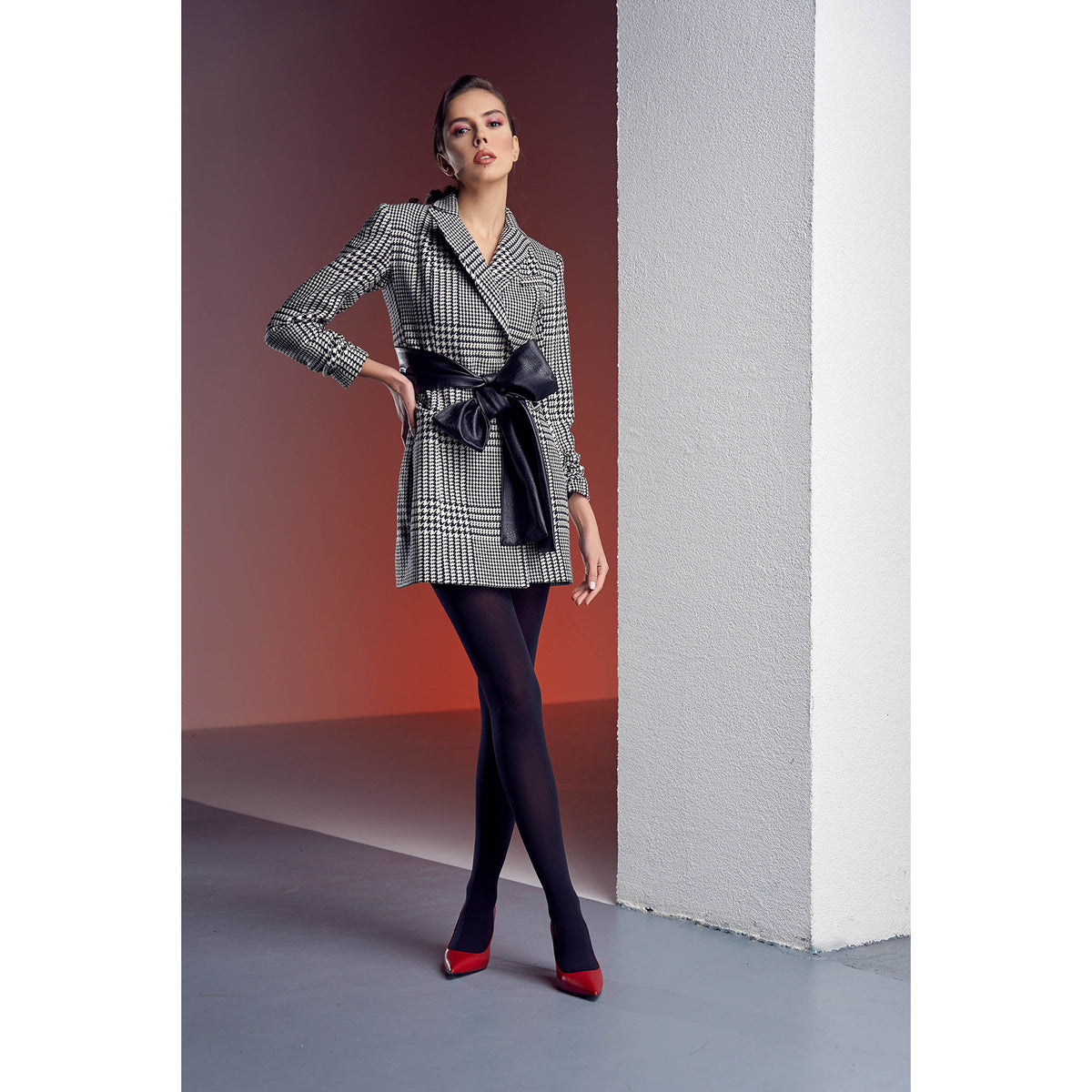 Woman wearing a checkered coat with a black bag against a white wall.