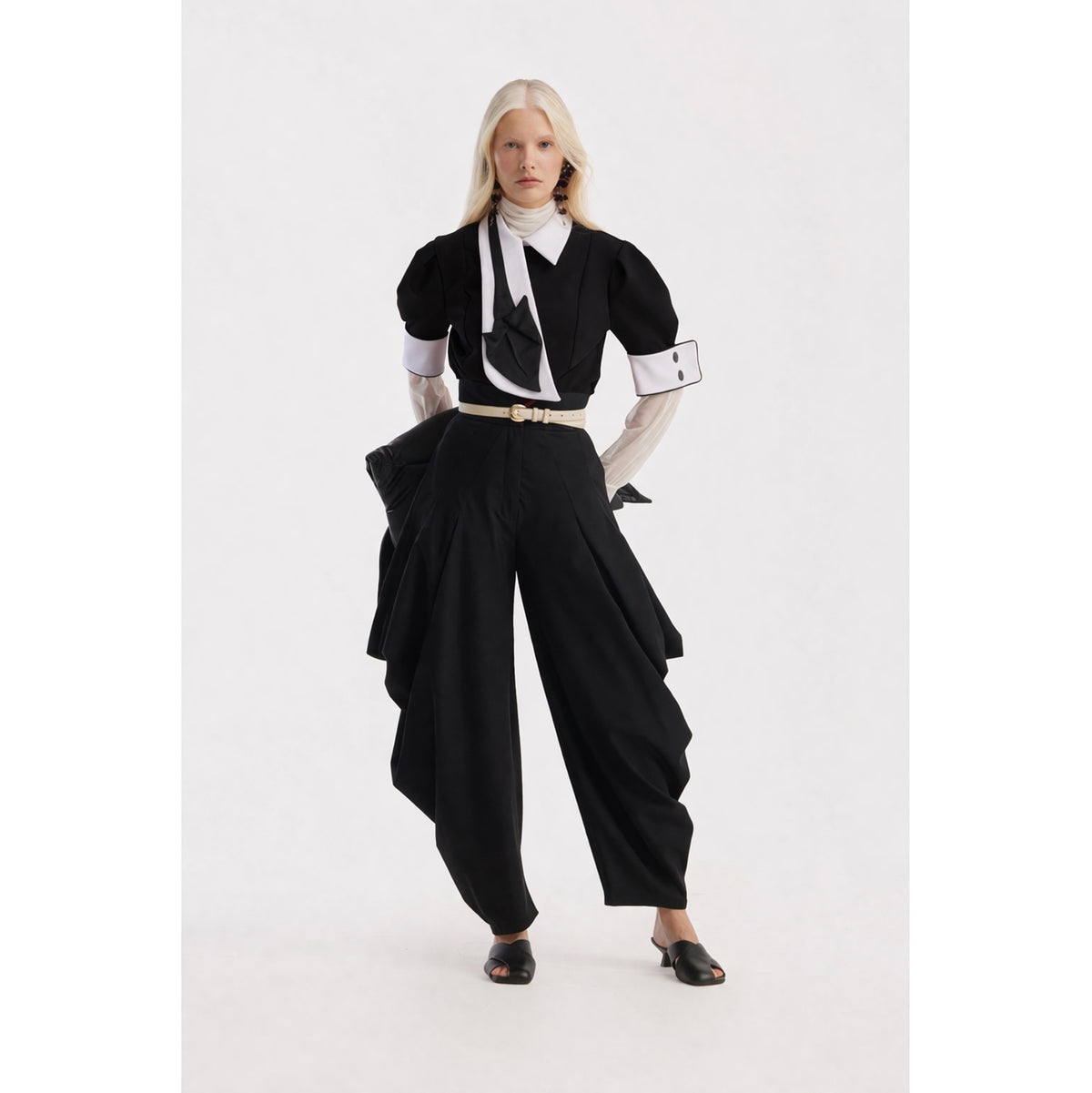 A person with long, light blonde hair wears a JULIA ALLERT Statement Blouse With White Collar And Cuffs in black, paired with wide-leg trousers, a white turtleneck, black mules, and a white belt against a plain background.