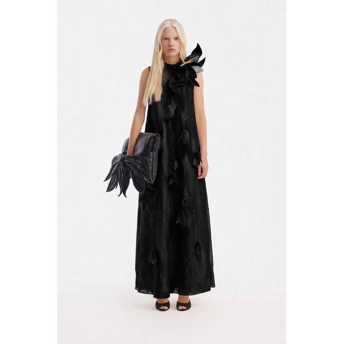 A woman with long blond hair wears the JULIA ALLERT Black A-Line Flower Maxi Dress—an elegant couture gown with petal details and a bold shoulder flower, styled with a large black bow clutch and black open-toe heels.