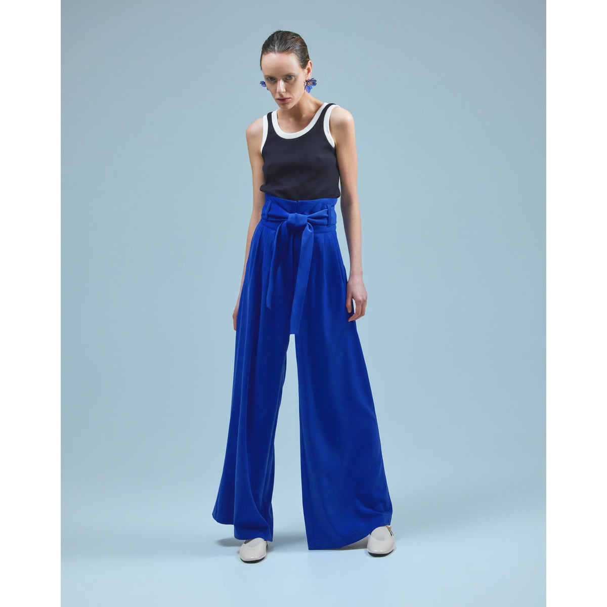 A woman poses against a light blue background in a sleeveless black and white top, JULIA ALLERT Blue High Waist Long Wide Leg Trousers with a bow at the waist, white shoes, and statement blue earrings.