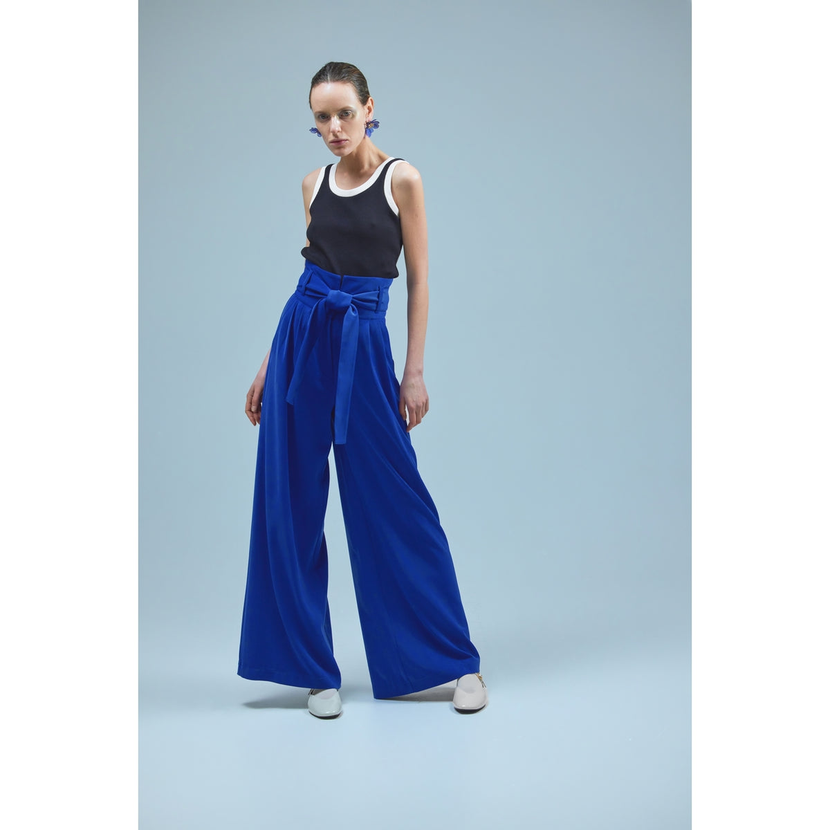 A woman with slicked-back hair and a serious expression stands against a light blue background wearing a black tank top with white trim, white shoes, and JULIA ALLERT Blue High Waist Long Wide Leg Trousers.