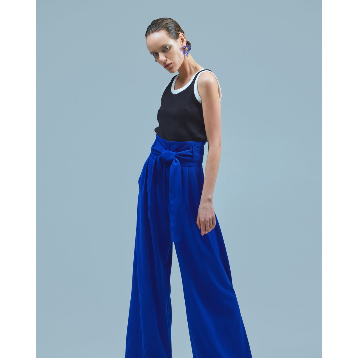 A woman poses against a light blue backdrop, wearing a sleeveless black top with white trim, JULIA ALLERT Blue High Waist Long Wide Leg Trousers with a matching belt, and blue statement earrings.