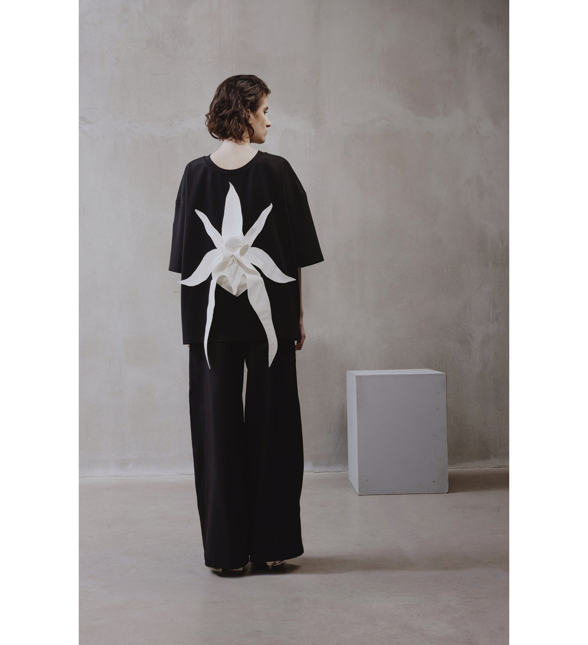 A woman stands indoors facing away, wearing the JULIA ALLERT Relaxed Fit Knit Top in black with wide pants and a sculptural floral appliqué on the back; a gray cube is nearby against a textured wall.