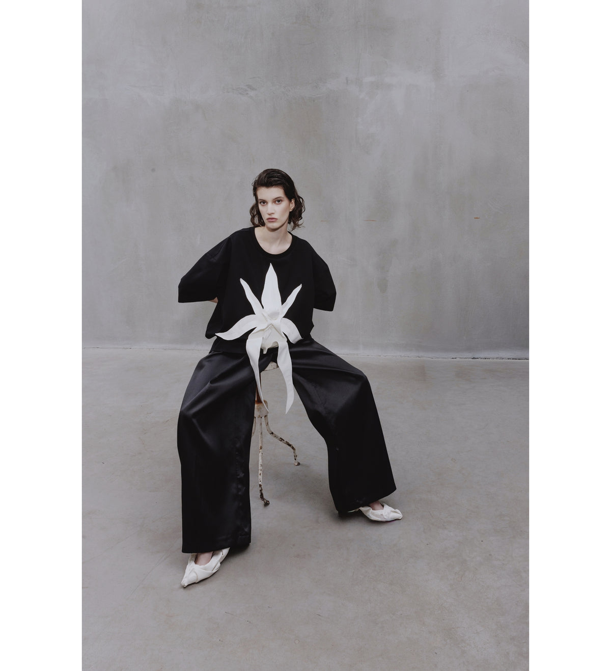 A person with short dark hair sits on a chair in a minimalist room, wearing JULIA ALLERT's Relaxed Fit Knit Top - Black with black wide-leg pants and white shoes, the top featuring a striking sculptural white floral appliqué.