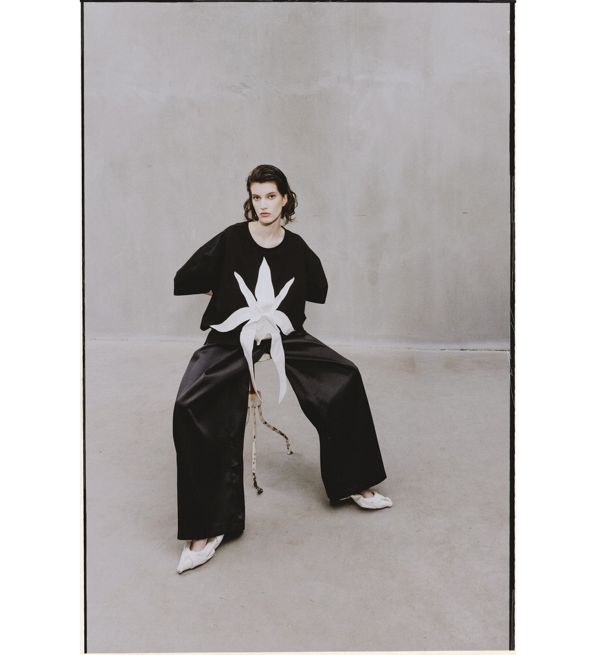 A person with short dark hair sits on a chair in a minimalist room, wearing wide black pants and white pointed shoes. Their top is the JULIA ALLERT Relaxed Fit Knit Top - Black, and they have a neutral expression.