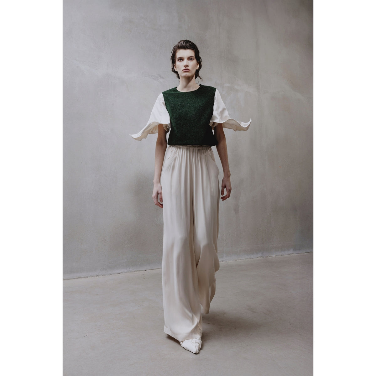 In a minimalist room, a person wears the JULIA ALLERT Green Lurex Knit Top With Adjustable Satin Sleeves over a white floor-length dress. The look is modern and elegant, set against soft lighting and a neutral background.