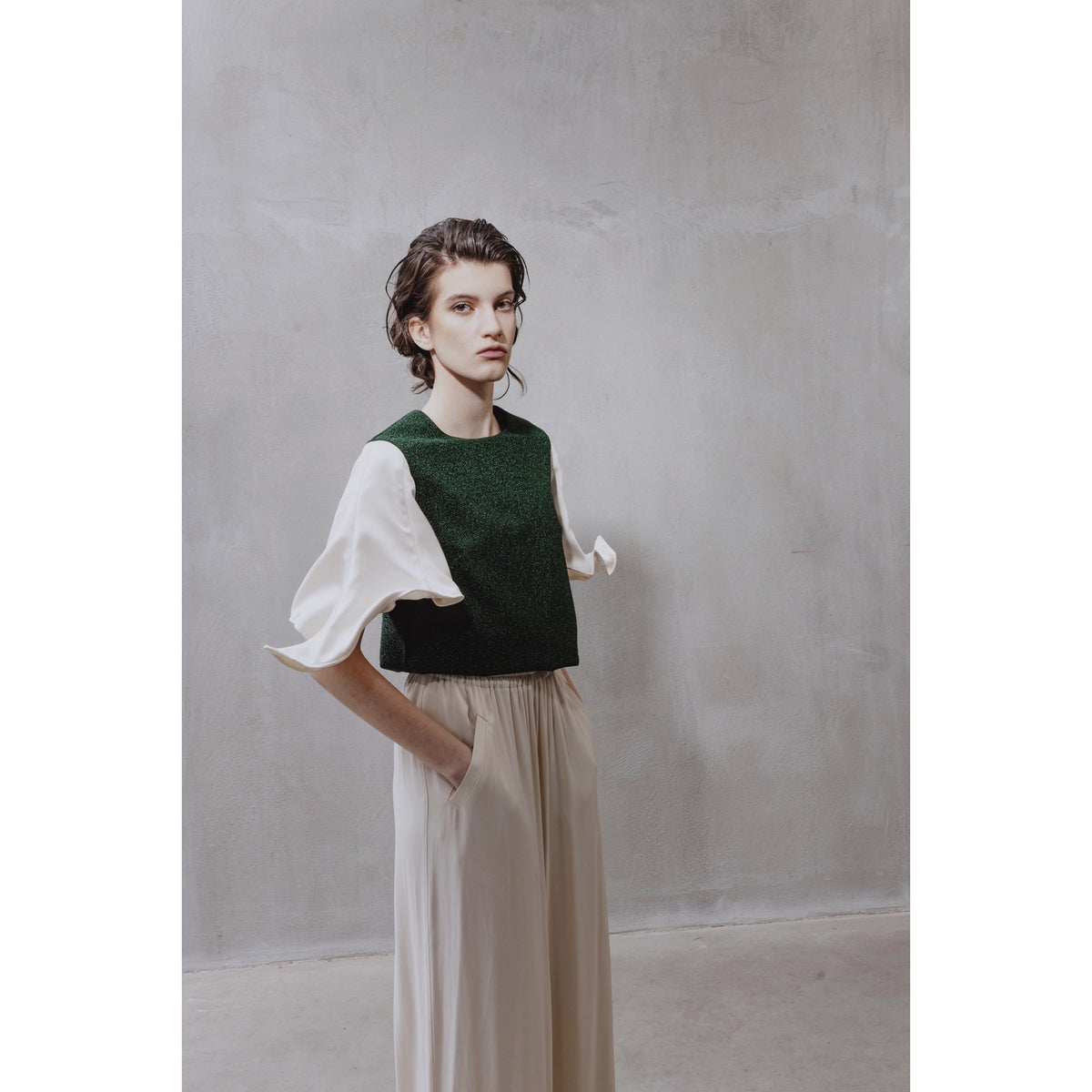 A person with short brown hair wears a white blouse, the JULIA ALLERT Green Lurex Knit Top With Adjustable Satin Sleeves, and cream high-waisted pants while standing against a light gray textured wall.
