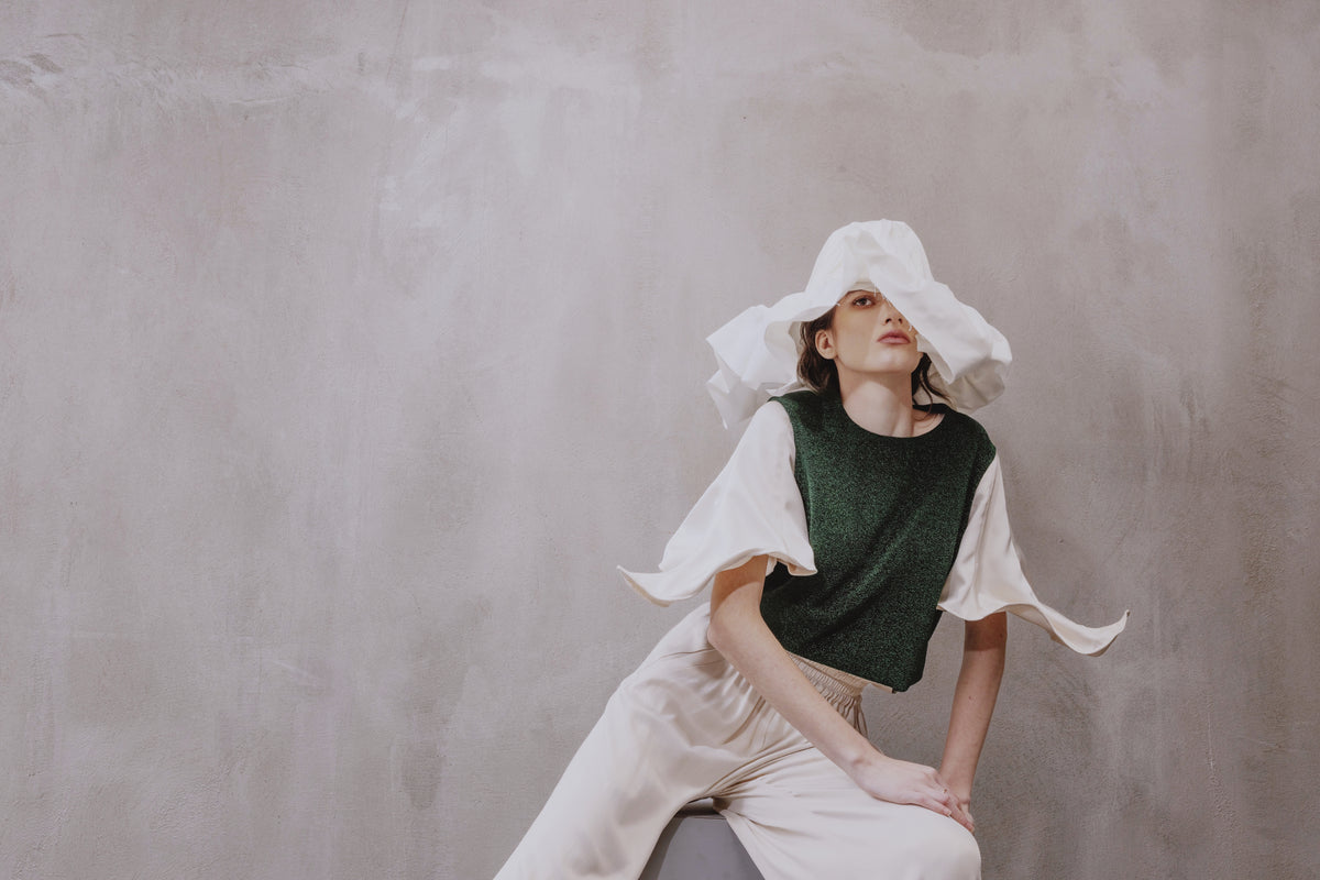 A woman poses by a textured gray wall in a JULIA ALLERT Green Lurex Knit Top With Adjustable Satin Sleeves, styled with a white wide-brim hat and loose white pants. Her neutral expression complements the modern, artistic look.