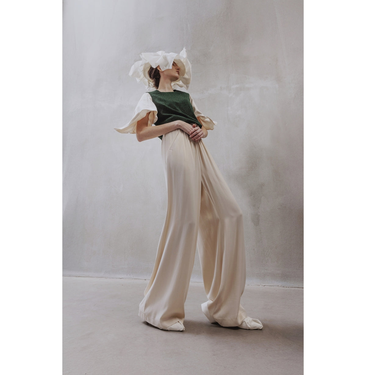 A person poses in a minimalist space, wearing wide-legged cream trousers and a JULIA ALLERT Green Lurex Knit Top With Adjustable Satin Sleeves, with a sheer white headpiece partially covering their face. The backdrop is a light gray textured wall.