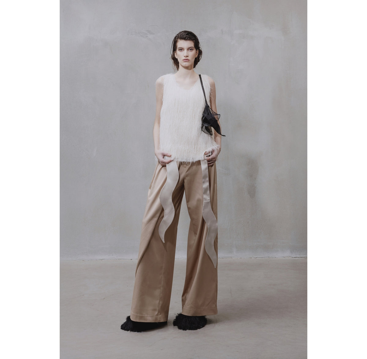 A woman stands against a textured gray wall, wearing a sleeveless white top, JULIA ALLERT High-Waist Wrap Trousers in beige, black ruffled shoes, and a small black shoulder bag.