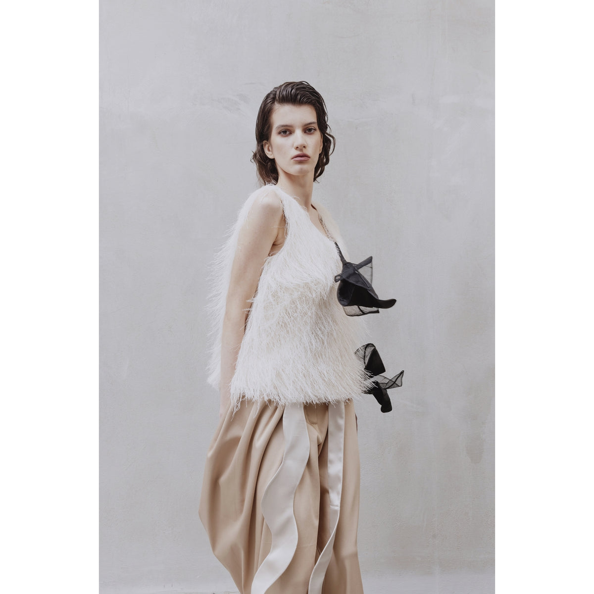 A woman with wet-look hair wears JULIA ALLERT High-Waist Wrap Trousers in beige, paired with a white furry sleeveless top featuring a large black bow, standing against a light grey textured wall for an elegant look.
