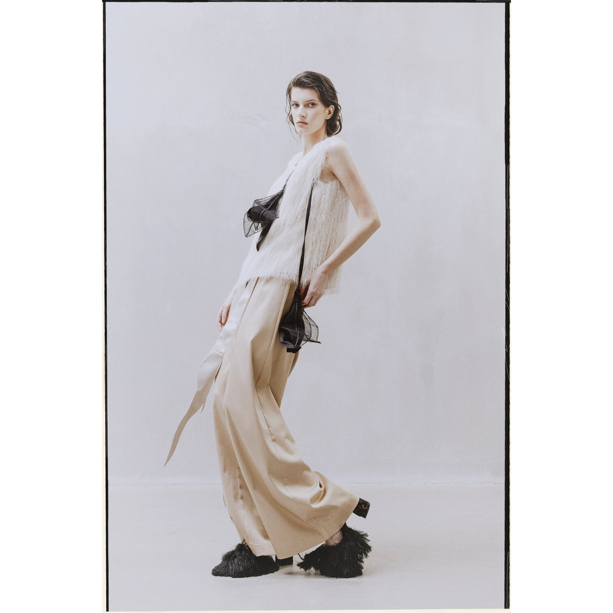 A person poses by a plain, light wall in a sleeveless white textured top, JULIA ALLERT High-Waist Wrap Trousers - Beige, black fluffy shoes, and holding a black sheer accessory for a modern, fashion-forward look.