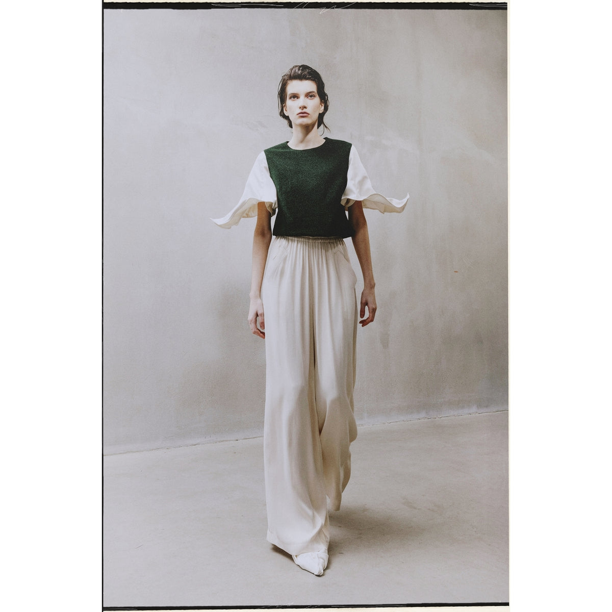 A person stands indoors in a JULIA ALLERT Green Lurex Knit Top With Adjustable Satin Sleeves, layered over a white blouse and long white skirt. The minimalist style is accentuated by the plain, neutral background.