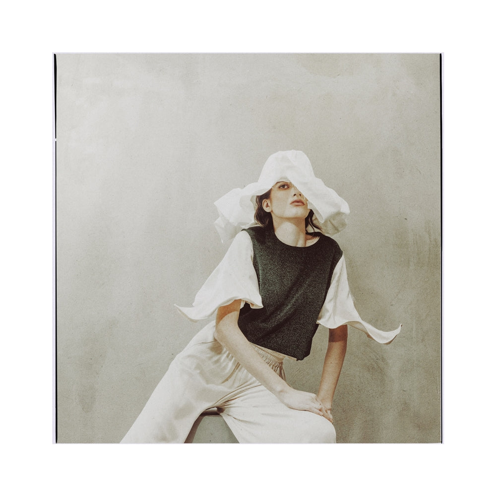 A woman poses against a neutral background in a JULIA ALLERT green lurex knit top with adjustable satin sleeves, paired with light-colored pants and a wide-brimmed white hat. One hand rests on her knee as she looks slightly upward.