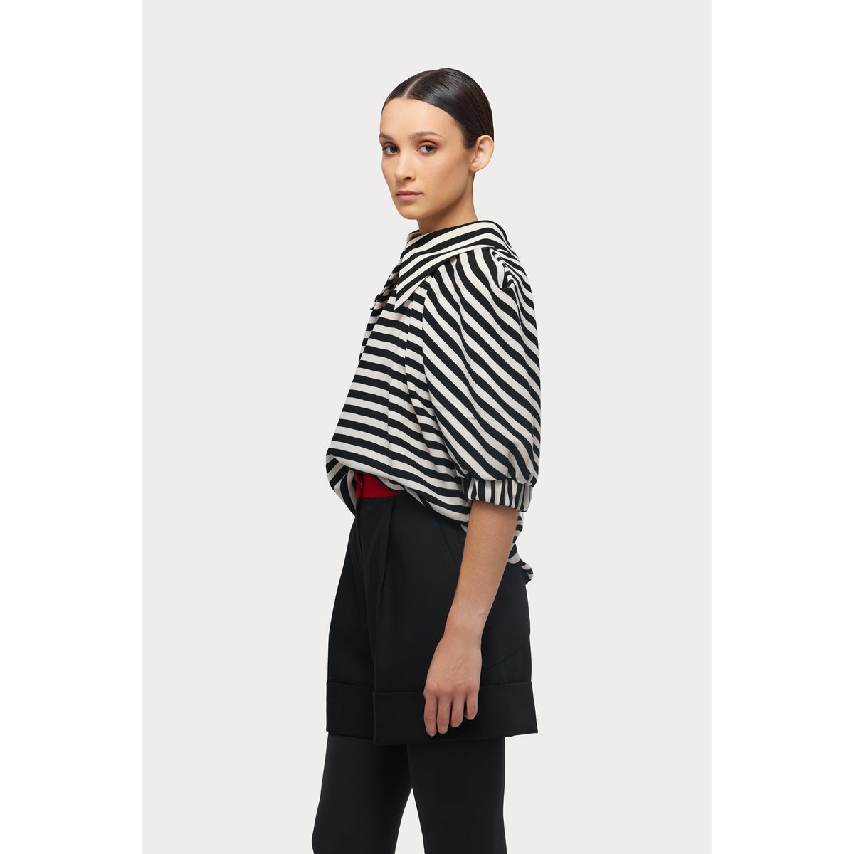 A person stands sideways in a JULIA ALLERT Designer Blouse With Black White Stripes, paired with black high-waisted shorts over black leggings against a plain white background.
