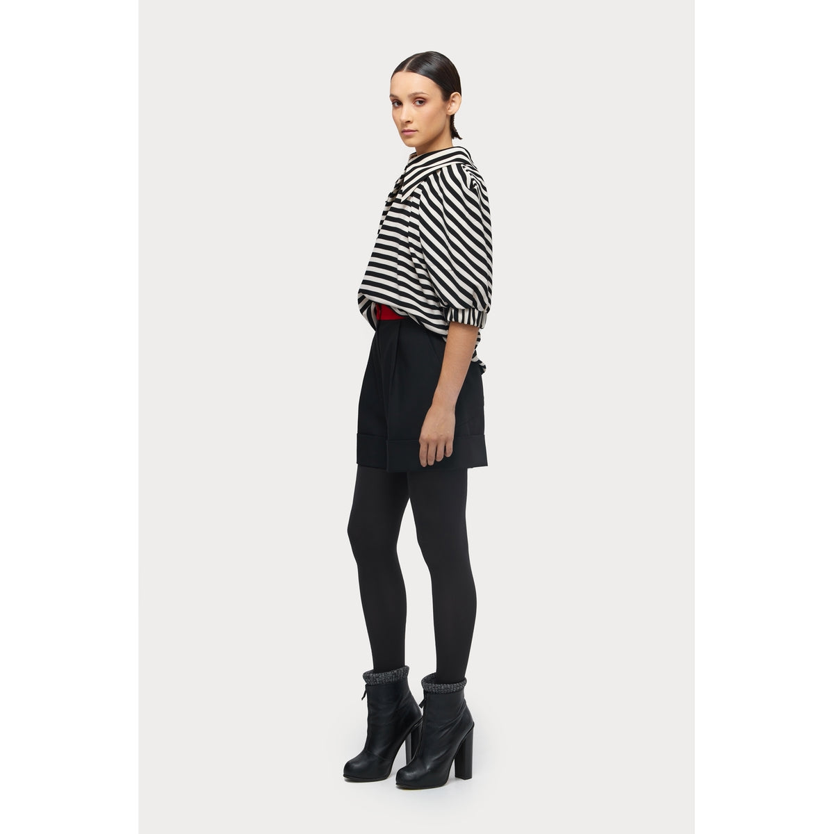 A woman stands in profile wearing a JULIA ALLERT Designer Blouse With Black White Stripes, black shorts, black tights, and black ankle boots against a plain white background.