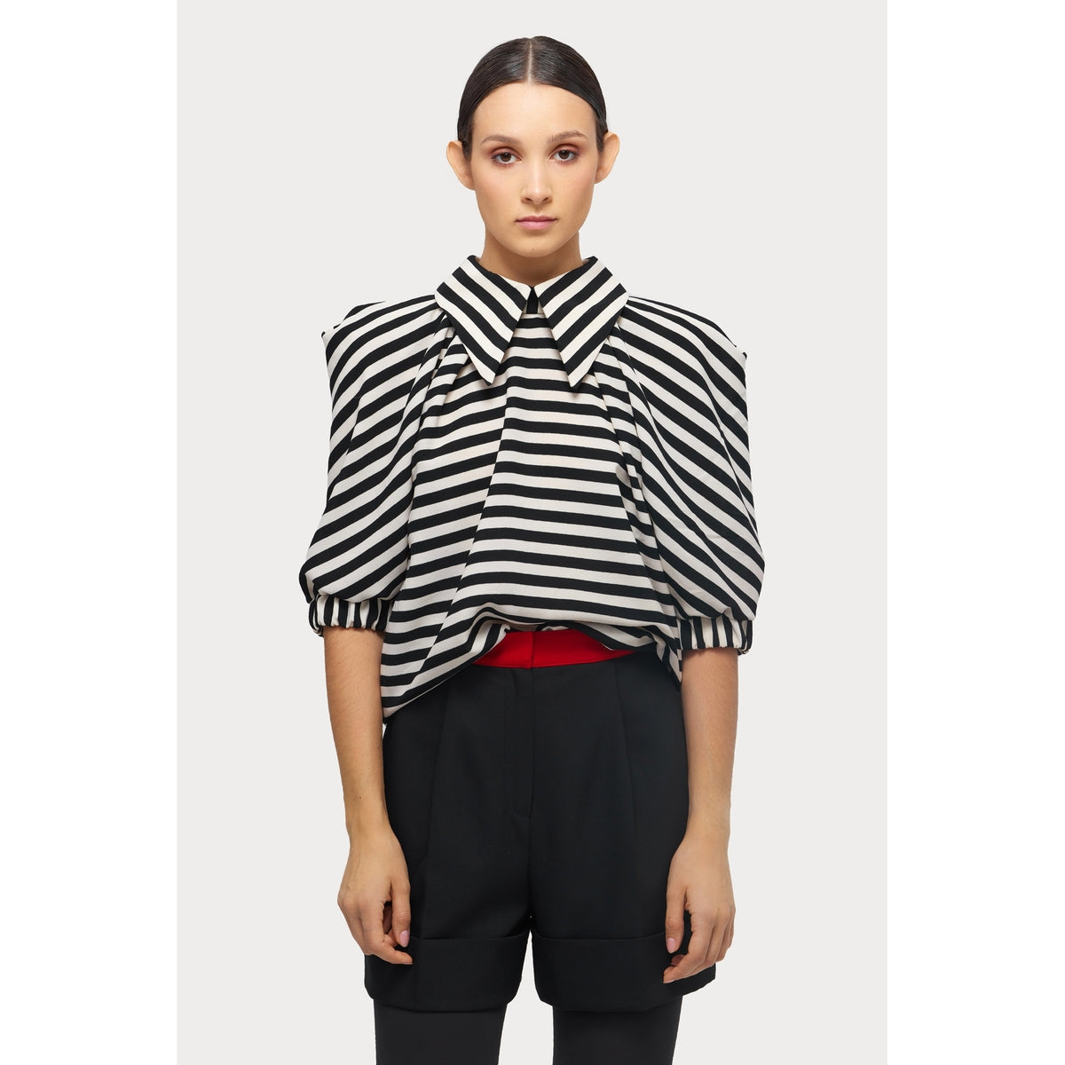 A person stands facing forward, wearing a JULIA ALLERT Designer Blouse With Black White Stripes featuring a wide collar, paired with high-waisted black shorts and a red belt. Their dark hair is neatly parted in the center.