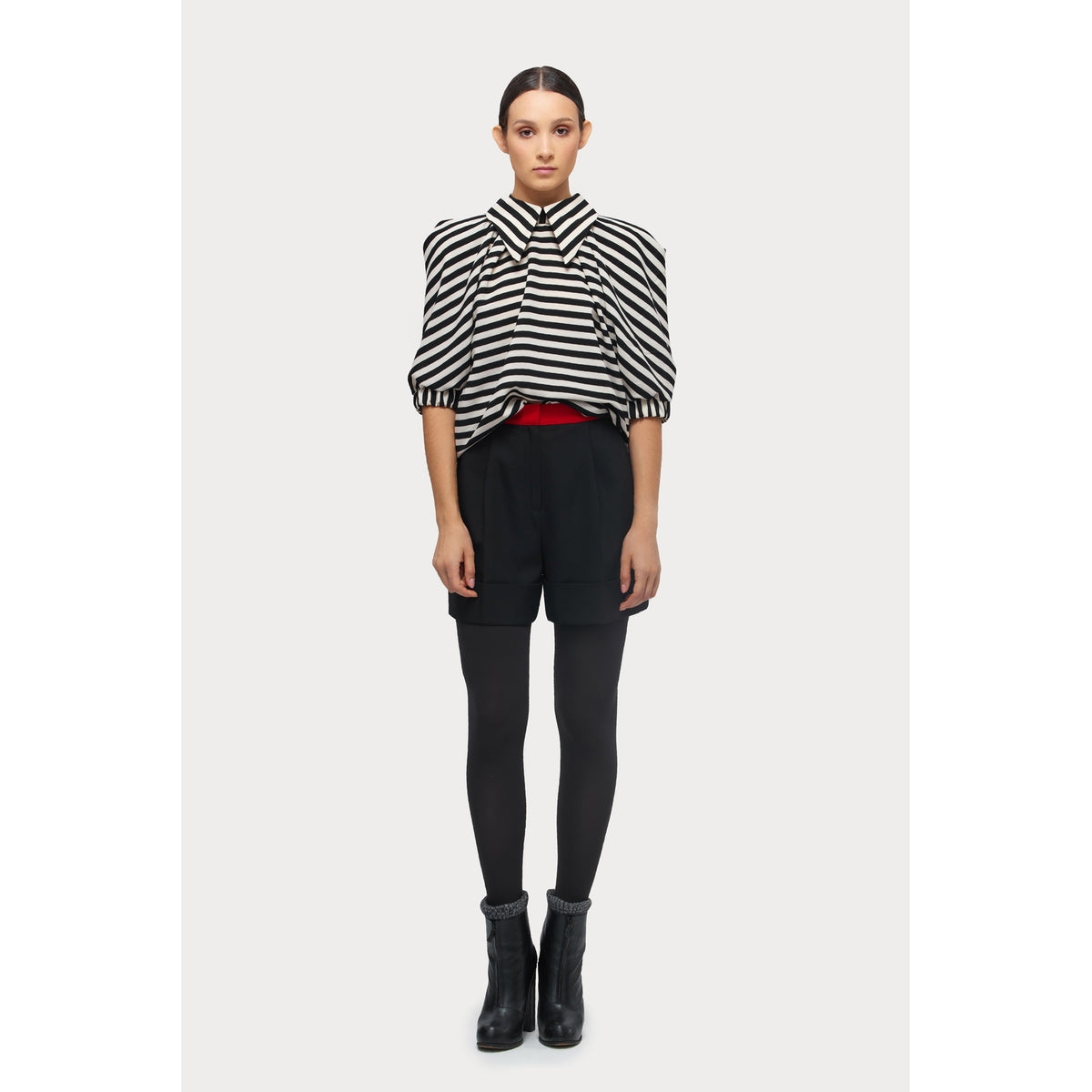A person stands facing forward against a plain white background, wearing a JULIA ALLERT Designer Blouse With Black White Stripes, high-waisted black shorts, black tights, black ankle boots, and a red belt.