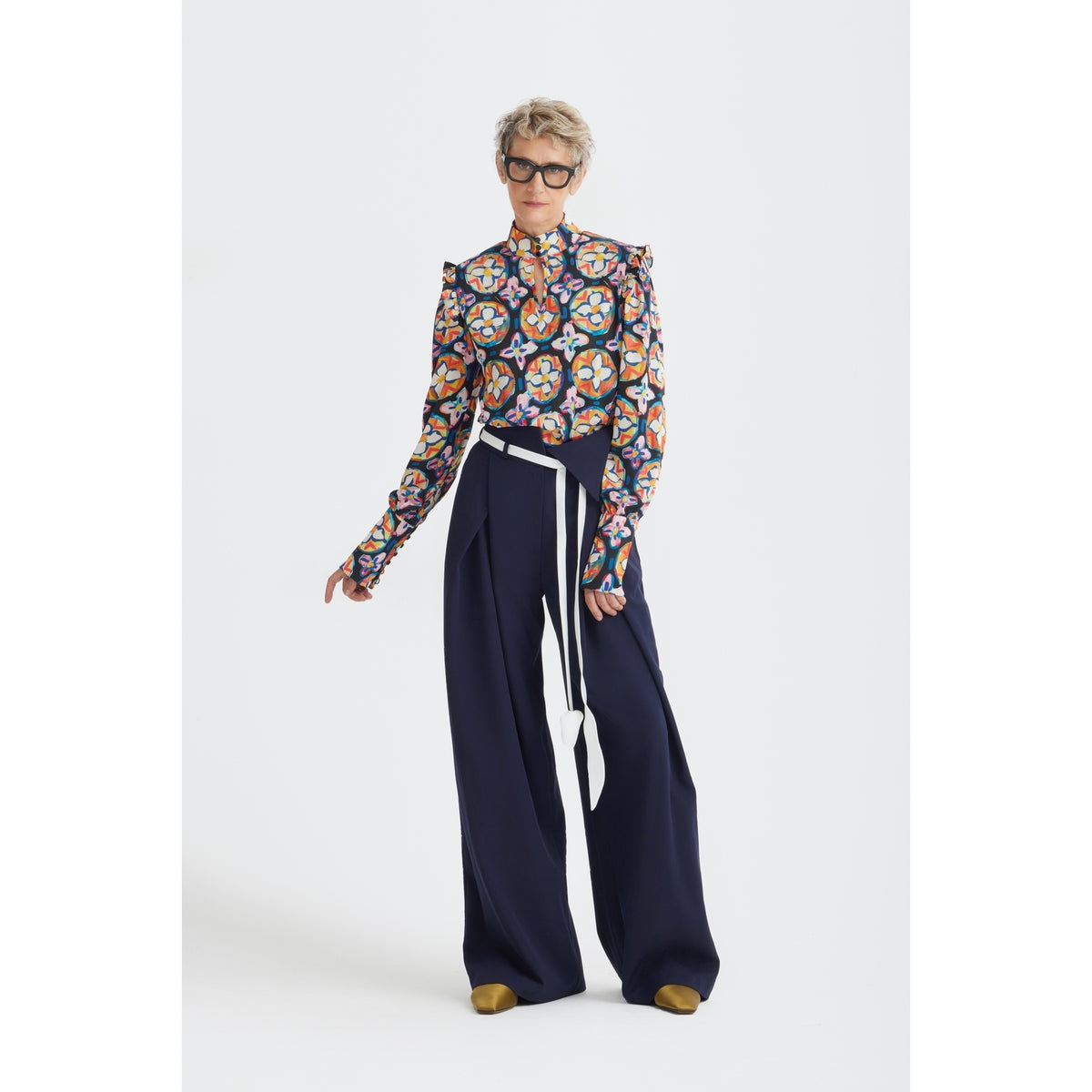 A person with short, light hair wears black glasses, a colorful patterned long-sleeve blouse, and JULIA ALLERT’s Fashion Forward Wide-Leg Pants &amp; Contrast Belt in navy, standing against a plain white background.