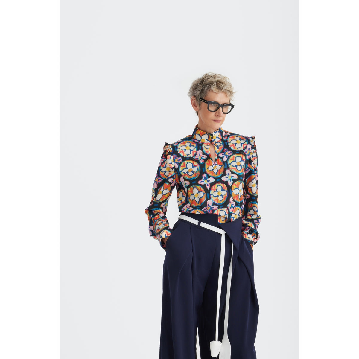 A person with short blond hair and black glasses wears JULIA ALLERT's Fashion Forward Wide-Leg Pants &amp; Contrast Belt in navy, paired with a colorful blouse, standing against a plain white background.