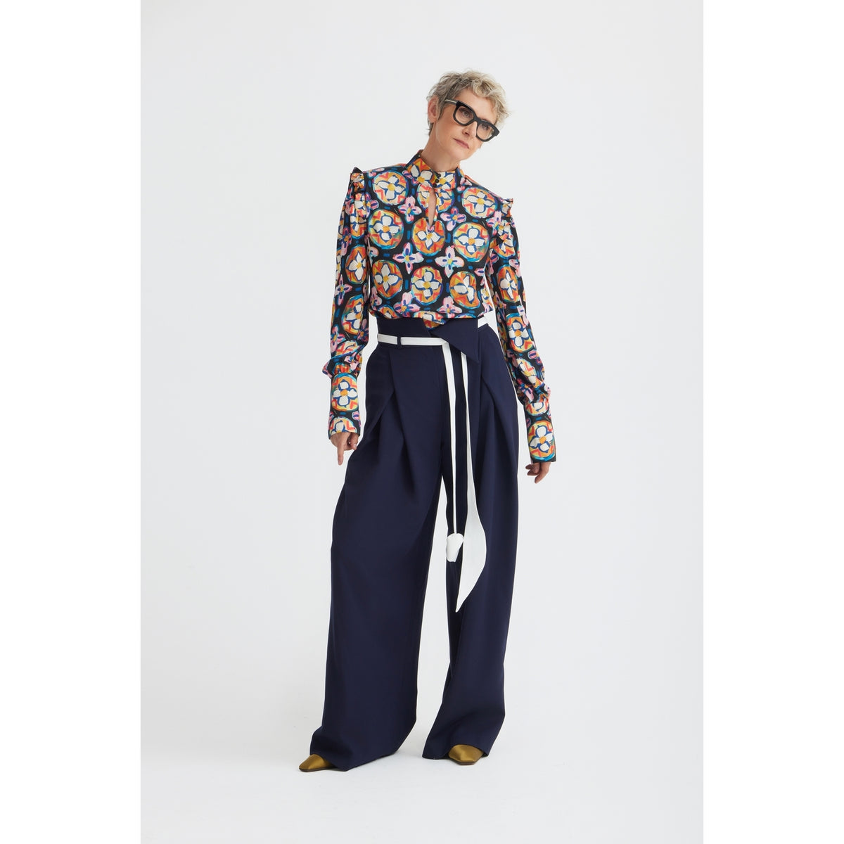 A person with short gray hair and glasses confidently poses in JULIA ALLERT’s Fashion Forward Wide-Leg Pants &amp; Contrast Belt - Navy, paired with a colorful patterned blouse, against a plain white background.