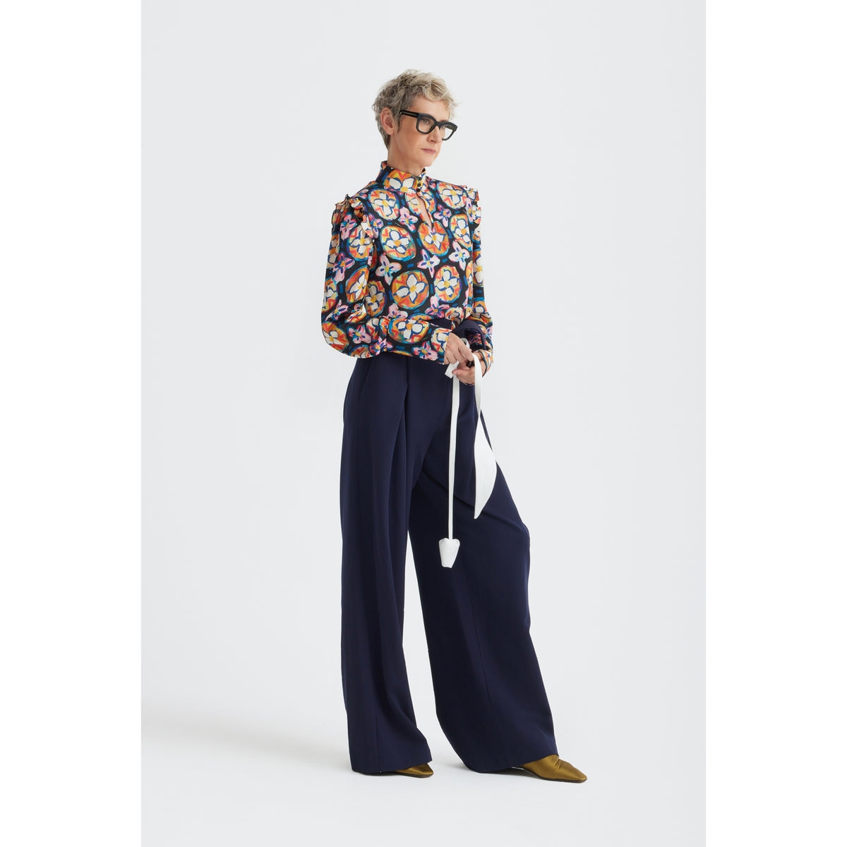 A person with short hair and glasses wears JULIA ALLERT’s Fashion Forward Wide-Leg Pants &amp; Contrast Belt in navy, styled with a floral blouse, gold shoes, and a white long-strap handbag against a plain white background.