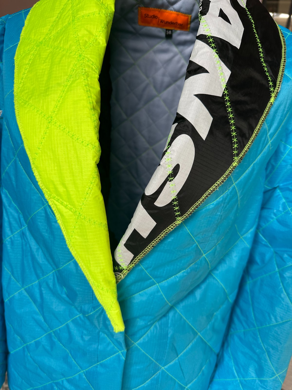 STUDIO WUNDERBAR Kite Classic: electric blue diamond-quilted maxi coat with neon green lapels and black-and-white graphic accents, women's and men's upcycled long coat, close-up detail, buy at DOORS NYC