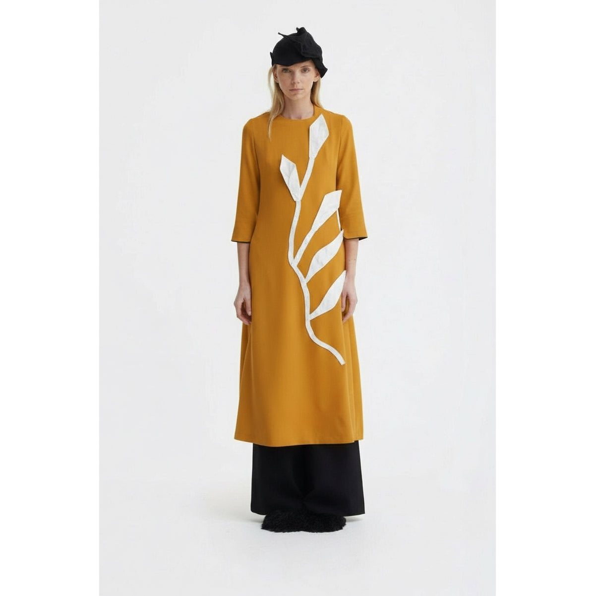 A person wearing the JULIA ALLERT Art-Inspired Semi-Fitted Three-Quarter Sleeves Dress in mustard, paired with black wide-leg pants, black shoes, and a black hat, stands against a plain white background.