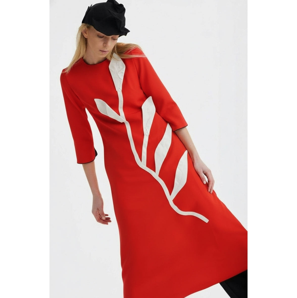 A woman in a JULIA ALLERT Art-Inspired Semi-Fitted Three-Quarter Sleeves Dress - Red poses against a plain white background, wearing a black hat. The dress features bright red fabric with white abstract leaf patterns and sculptural appliqué.