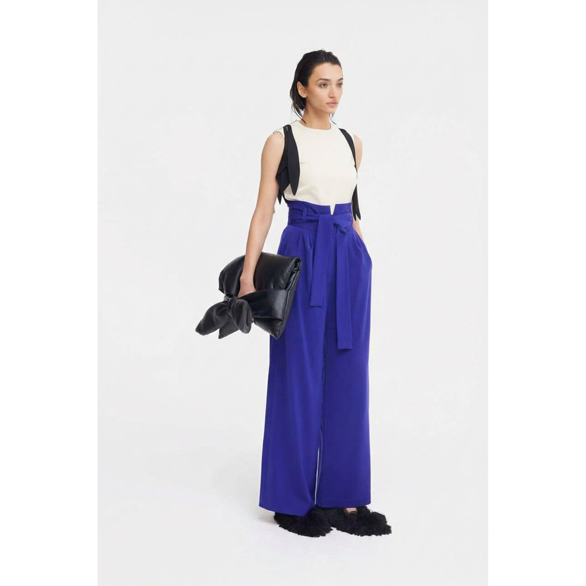 A woman wears JULIA ALLERT’s Designer Fitted Sleeveless Top with Floral Accent in ivory, paired with high-waisted royal blue wide-leg trousers and black fuzzy shoes, holding a large black bow clutch against a plain white background.