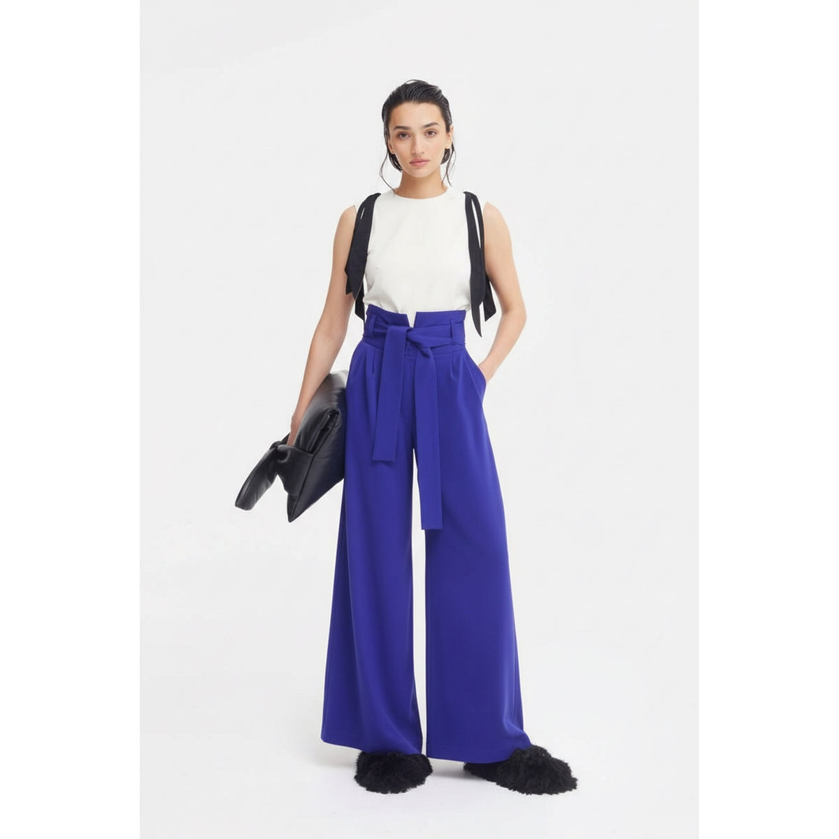 A woman stands against a white background, wearing the JULIA ALLERT Designer Fitted Sleeveless Top with floral accents in ivory, paired with high-waisted wide-leg cobalt blue pants, black furry shoes, and holding a large black handbag.