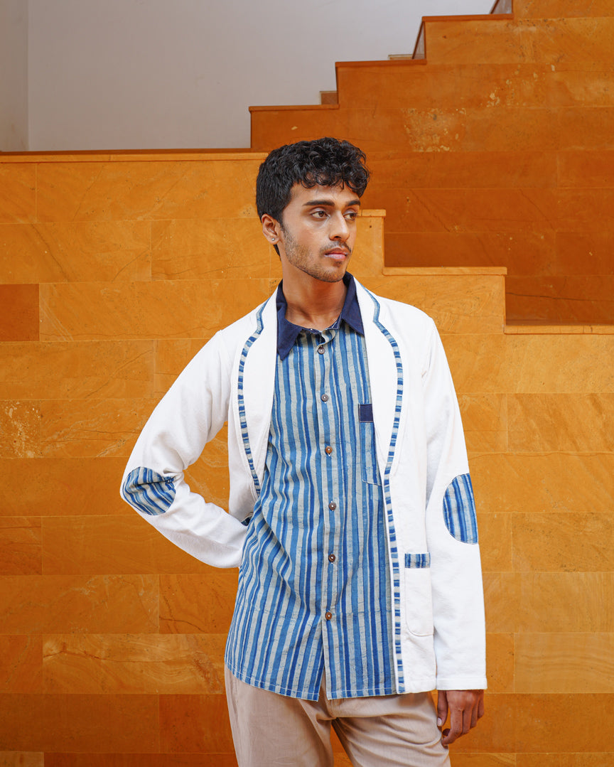 A young man stands before orange-brown stairs, wearing light tan pants, a blue and white striped shirt, and the JIWYA Baadal Unisex Jacket in White Colour with Indigo Elements. He looks to the side with one hand on his hip.