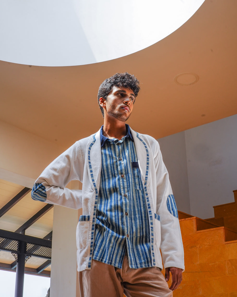 A young man with short curly hair stands indoors by a staircase, wearing a blue striped shirt and the Baadal Unisex Jacket in White Colour and Indigo Elements by JIWYA. Natural light pours in from a large circular skylight above.
