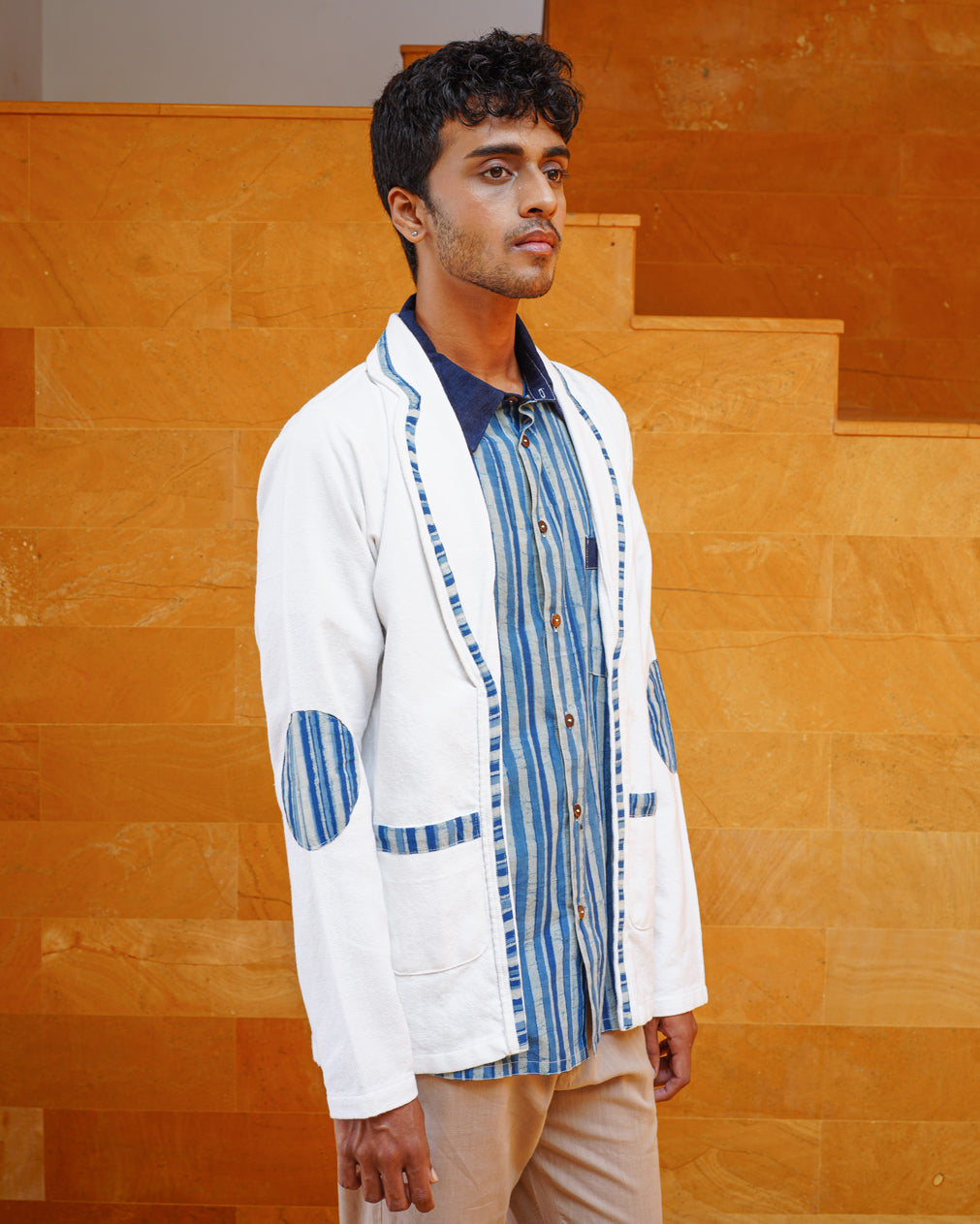 A young man with short dark hair stands before ochre stairs, wearing the JIWYA Baadal Unisex Jacket in White Colour and Indigo Elements over a blue striped shirt and beige pants.