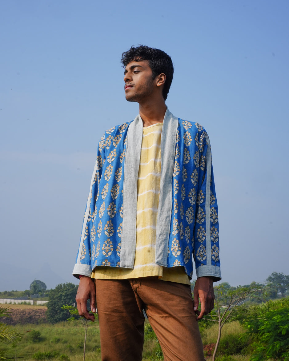 A young man stands outdoors in the JIWYA Nadi Reversible Unisex Jacket over a yellow shirt and brown pants, gazing confidently to the side with fields, greenery, and a clear sky behind him.