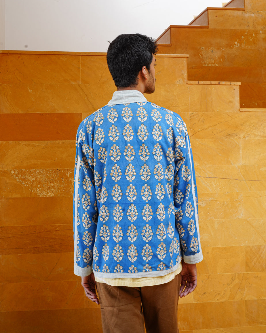 A person with short dark hair faces away, wearing the JIWYA Nadi Reversible Unisex Jacket with white floral patterns on sustainable cotton and brown pants, standing before yellow stone stairs and a matching wall.