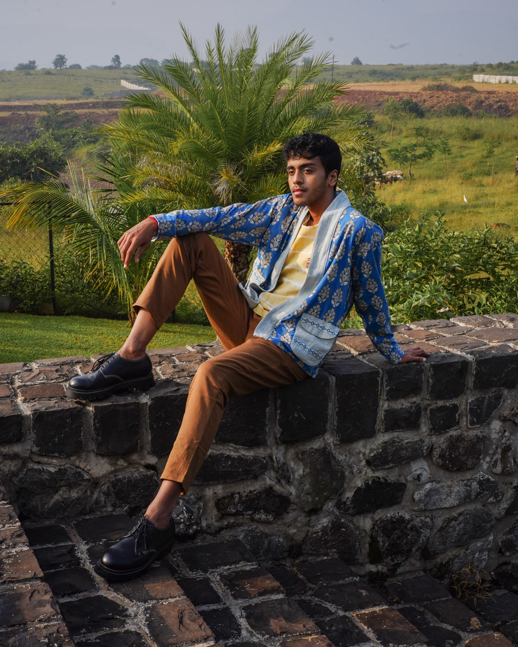 A man wears the JIWYA Nadi Reversible Unisex Jacket over a yellow shirt and brown pants as he sits on a stone wall outdoors, surrounded by green grass, palm trees, and a scenic countryside under a cloudy sky.