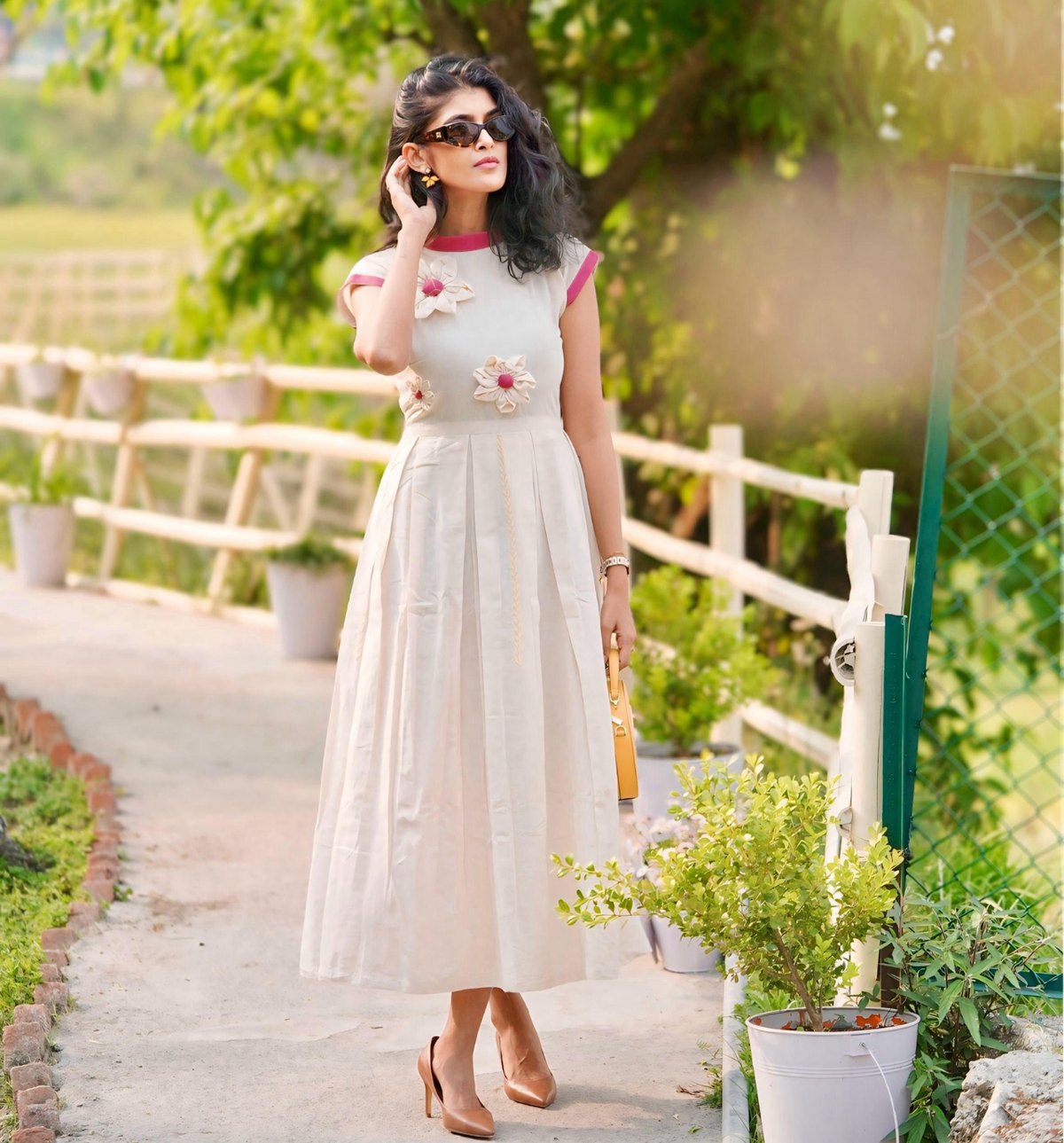 A woman wearing the cream Papiha Midi Dress by JIWYA, featuring upcycled 3D flowers, stands on a garden path with a purse, sunglasses, and heels, surrounded by greenery and potted plants.