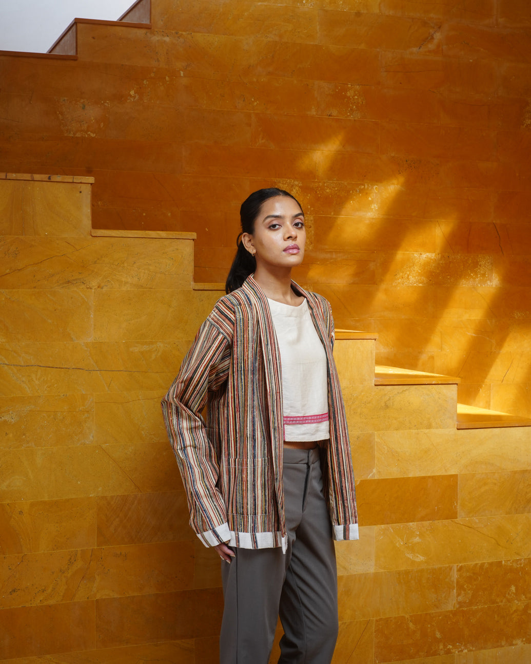 A woman stands on a golden-brown stone staircase, confidently wearing the JIWYA Surajmukhi Unisex Jacket, made from rainfed cotton, over a white top and gray pants as warm sunlight fills the scene.
