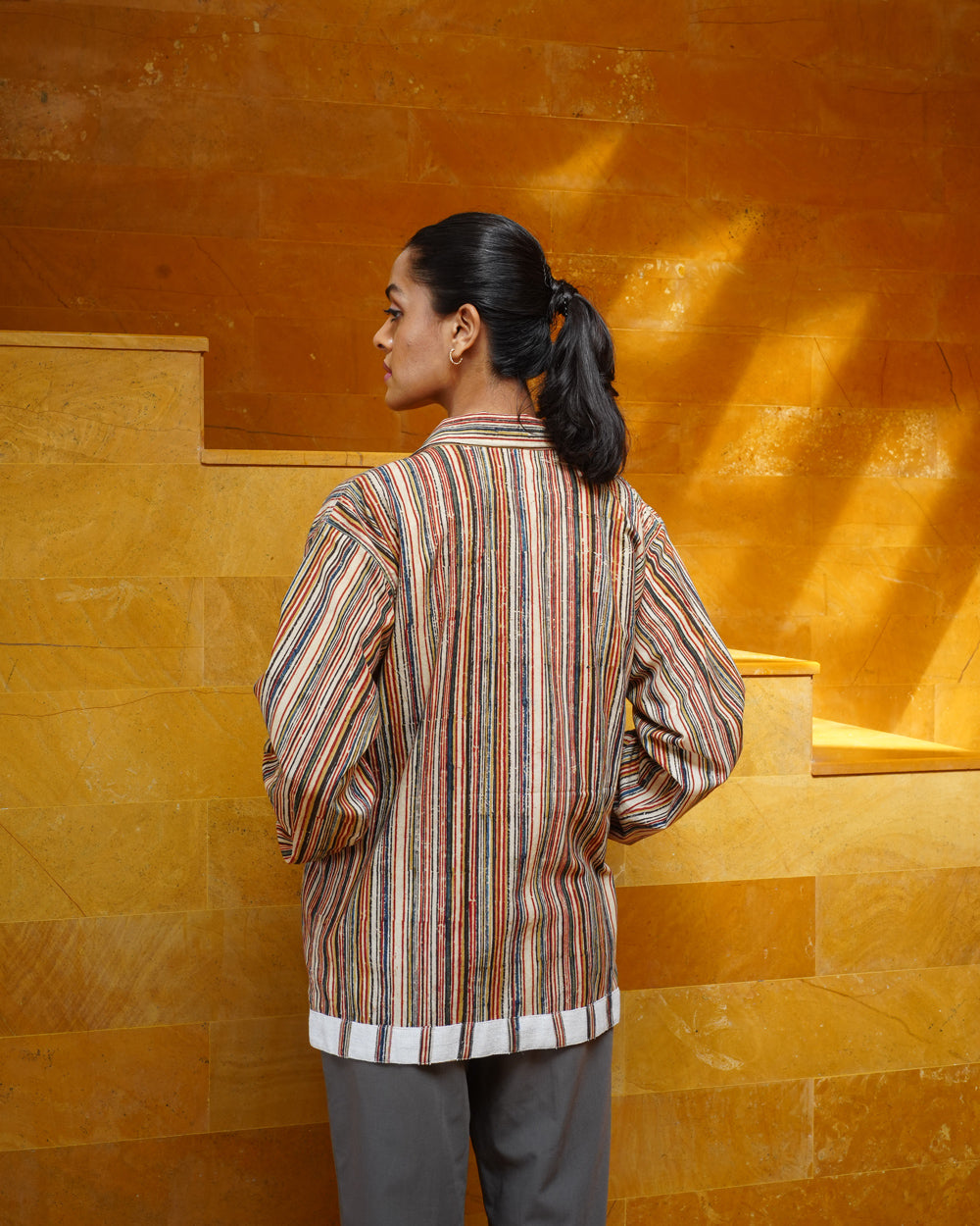 A woman with dark hair in a ponytail stands on warmly lit stone steps, wearing the JIWYA Surajmukhi Unisex Jacket, featuring vertical multicolored stripes and crafted from rainfed cotton.