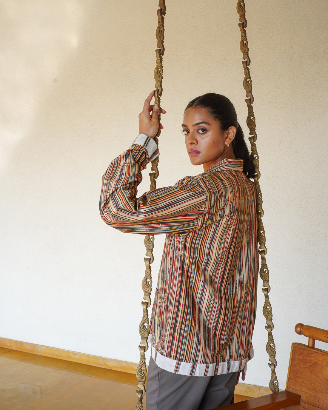 A person with long dark hair wears the JIWYA Surajmukhi Unisex Jacket, made from rainfed cotton, and gray pants. They stand sideways on a wooden swing with metal chains, looking over their shoulder before a plain beige wall.